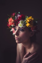 A person is adorned with a vibrant floral crown composed of various flowers in shades of red, white, yellow, and green. The lighting is soft and moody, casting gentle shadows across the face, conveying a contemplative or serene expression.