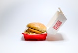 A sesame seed hamburger bun encases crispy fried chicken in a red and white fast food box, slightly open and tilted, with branding text visible on the lid. The box rests against a plain white background, emphasizing the simplicity and cleanliness of the presentation.