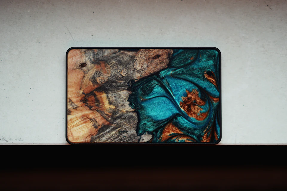 The image shows a rectangular, black-framed object with a vibrant blue and gold design on its surface. The frame is positioned against a white background, which contrasts with the colorful artwork. The edges of the frame are slightly frayed, adding a touch of rustic charm to the overall presentation.