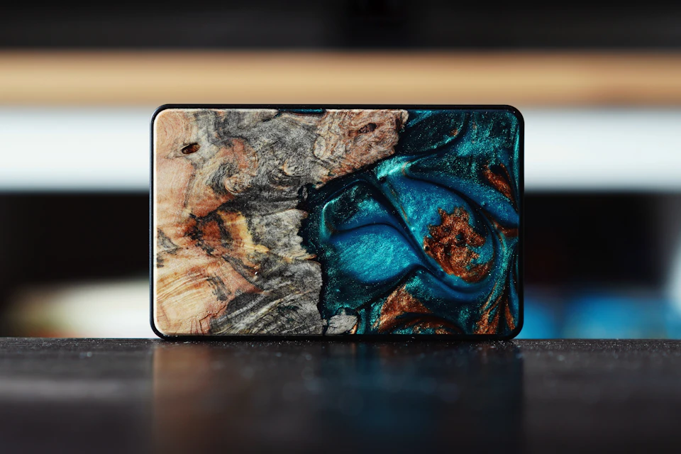 The image shows a rectangular, black plastic wallet resting on a wooden surface. The wallet has a unique design featuring blue and brown hues, with a pattern resembling a tree or plant-like structure. The wallet is positioned slightly to the left of center in the frame, allowing for a clear view of its details.