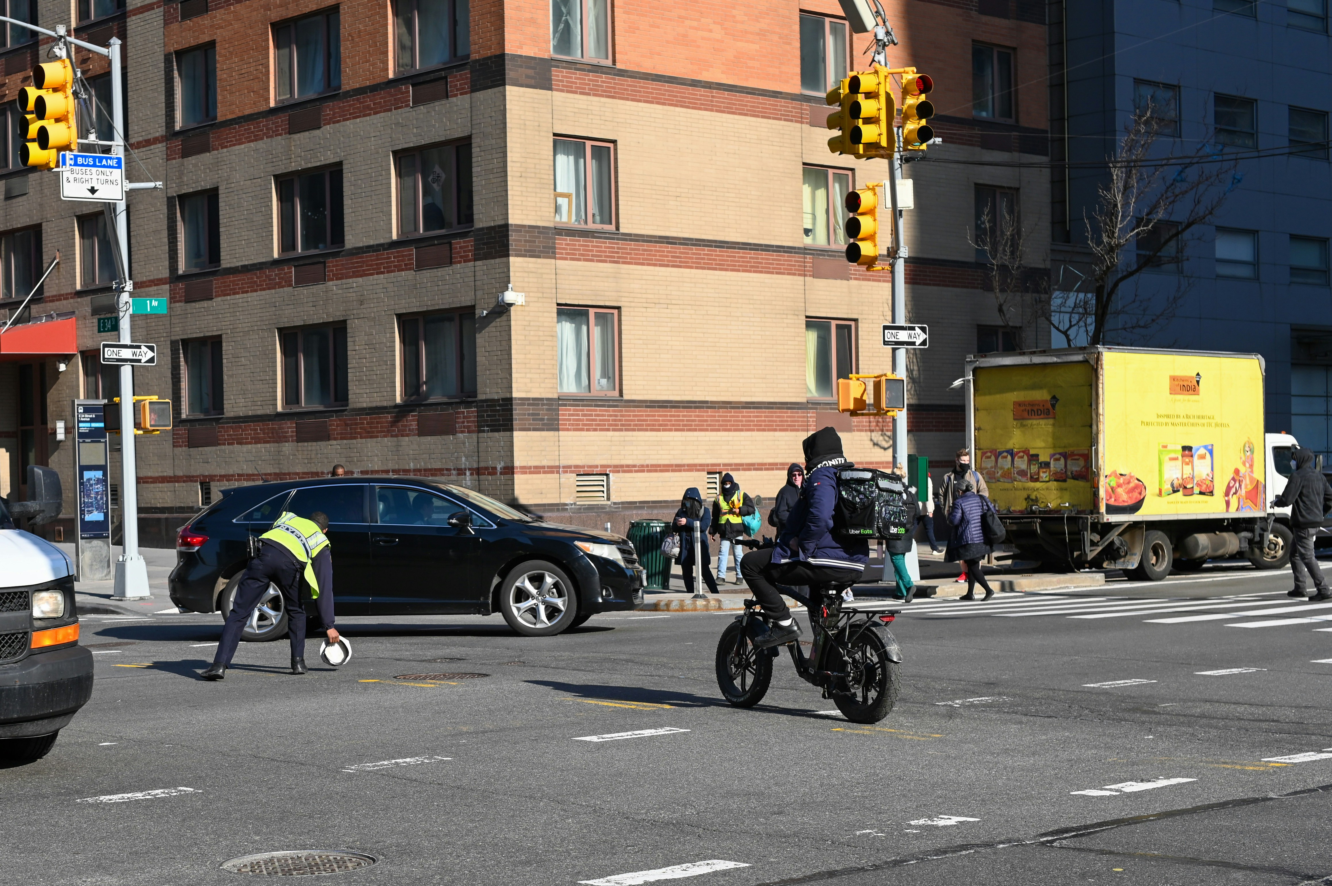 A busy urban street corner with a multi-story brick building in the background. A traffic officer is directing or managing traffic near a stopped black car and a delivery truck is parked on the side with bright yellow advertising. Several pedestrians in winter clothing are waiting at the corner, and a person on a bicycle rides through the intersection.