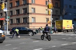 A busy urban street corner with a multi-story brick building in the background. A traffic officer is directing or managing traffic near a stopped black car and a delivery truck is parked on the side with bright yellow advertising. Several pedestrians in winter clothing are waiting at the corner, and a person on a bicycle rides through the intersection.