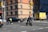 A busy urban street corner with a multi-story brick building in the background. A traffic officer is directing or managing traffic near a stopped black car and a delivery truck is parked on the side with bright yellow advertising. Several pedestrians in winter clothing are waiting at the corner, and a person on a bicycle rides through the intersection.