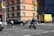 A busy urban street corner with a multi-story brick building in the background. A traffic officer is directing or managing traffic near a stopped black car and a delivery truck is parked on the side with bright yellow advertising. Several pedestrians in winter clothing are waiting at the corner, and a person on a bicycle rides through the intersection.
