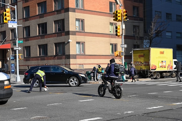 A busy urban street corner with a multi-story brick building in the background. A traffic officer is directing or managing traffic near a stopped black car and a delivery truck is parked on the side with bright yellow advertising. Several pedestrians in winter clothing are waiting at the corner, and a person on a bicycle rides through the intersection.