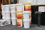 A stack of large white plastic buckets with yellow lids is prominently displayed. Some buckets have labels for a brand called 'Mr. Pickle,' advertising pickles and sauerkraut. Nearby is an orange box labeled 'Jumbo Yams.' The background shows a storefront with menus and a sign labeled 'Sprinkler Siamese' next to a fire hydrant connection.