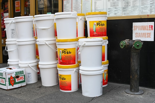 A stack of large white plastic buckets with yellow lids is prominently displayed. Some buckets have labels for a brand called 'Mr. Pickle,' advertising pickles and sauerkraut. Nearby is an orange box labeled 'Jumbo Yams.' The background shows a storefront with menus and a sign labeled 'Sprinkler Siamese' next to a fire hydrant connection.