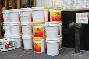 A stack of large white plastic buckets with yellow lids is prominently displayed. Some buckets have labels for a brand called 'Mr. Pickle,' advertising pickles and sauerkraut. Nearby is an orange box labeled 'Jumbo Yams.' The background shows a storefront with menus and a sign labeled 'Sprinkler Siamese' next to a fire hydrant connection.