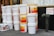 A stack of large white plastic buckets with yellow lids is prominently displayed. Some buckets have labels for a brand called 'Mr. Pickle,' advertising pickles and sauerkraut. Nearby is an orange box labeled 'Jumbo Yams.' The background shows a storefront with menus and a sign labeled 'Sprinkler Siamese' next to a fire hydrant connection.
