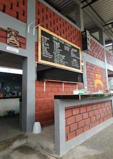 An outdoor cafe area with red and gray brick walls and a chalkboard menu. The space includes a counter with dishware and cleaning supplies. A speaker is mounted on the wall, and various kitchen utensils are visible in the background.