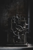 An old, industrial machine sits in a dimly lit, possibly abandoned factory setting. The machinery appears rustic and worn, with a large gear wheel and several mechanical components visible.