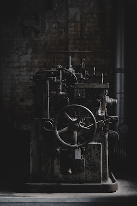 An old, industrial machine sits in a dimly lit, possibly abandoned factory setting. The machinery appears rustic and worn, with a large gear wheel and several mechanical components visible.