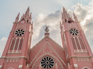 A grand Gothic-style church with two tall, intricately designed pink spires topped with crosses. The facade features ornate detailing with round and arched windows and a central statue of a religious figure. The backdrop includes a cloudy sky with sunlight peeking through.
