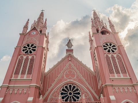 A grand Gothic-style church with two tall, intricately designed pink spires topped with crosses. The facade features ornate detailing with round and arched windows and a central statue of a religious figure. The backdrop includes a cloudy sky with sunlight peeking through.