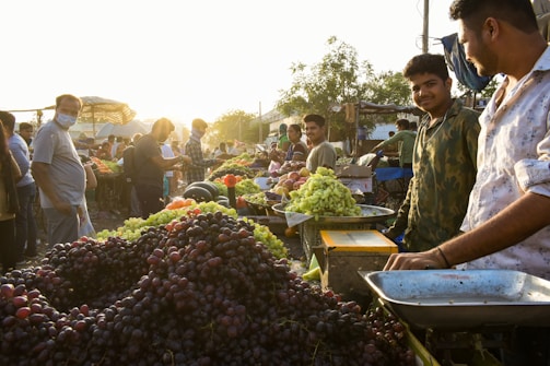 A bustling outdoor market scene featuring vendors selling a variety of fruits, including grapes and apples. Several people, including masked individuals, are seen interacting and purchasing produce as the sun sets in the background, casting a warm glow over the scene.