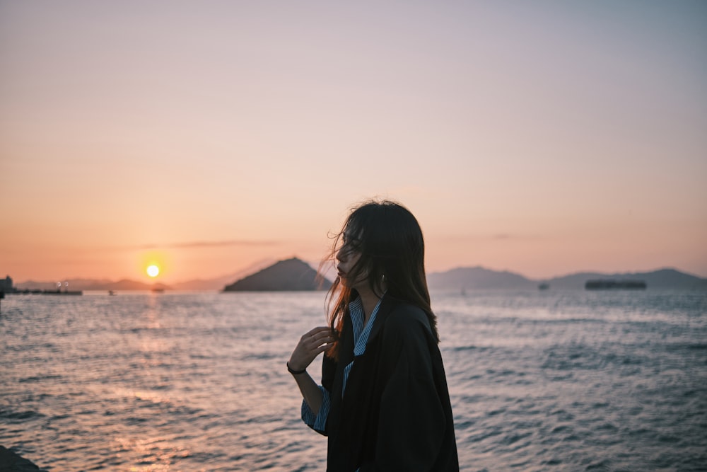 Frau im schwarzen Mantel steht bei Sonnenuntergang am Strand