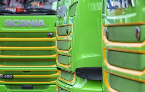 Close-up view of the front grills of two bright green trucks with yellow trim. The trucks have the brand name prominently displayed in large letters above the grills. The design features bold lines and a glossy finish.