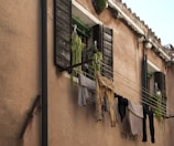 An exterior view of an old building with two windows. Laundry is hanging on clotheslines strung between the windows, including pants and shirts in muted colors. Wooden shutters, some hanging plants, and a stone wall contribute to a rustic, charming atmosphere.