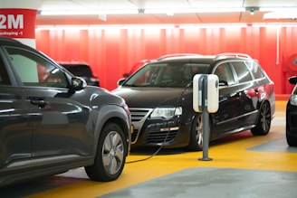 A parking garage with an electric car charging station. Two dark-colored cars are parked side by side, with one connected to the charging unit. The floor is marked with bright yellow and gray sections, and the walls are painted red. Overhead fluorescent lights illuminate the space, and a sign in the background marks the level as -1.