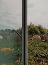The scene displays a split view through a glass window, dividing a rustic garden with a lush green yard filled with leafy plants and flowers. Two wooden sheds or small buildings can be seen on each side, surrounded by a wooden fence. The sky above is overcast, casting a muted light over the landscape.
