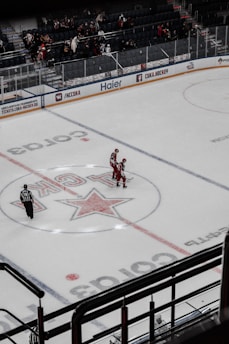 An ice hockey rink features two players in red uniforms celebrating on the rink. A referee is nearby, and there are spectators in the stands. The ice is marked with logos and center lines.
