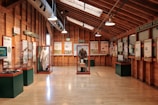 A museum exhibit room with wooden walls and ceiling, featuring various displays and informational panels. Displays include historical artifacts such as clothing and other cultural items protected in glass cases. Signs indicate different themes or topics, with visible text such as 'Columbia River Gorge' and 'Epic Northwest Flights.' The room is well-lit with natural and artificial lighting.