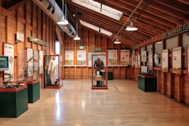 A museum exhibit room with wooden walls and ceiling, featuring various displays and informational panels. Displays include historical artifacts such as clothing and other cultural items protected in glass cases. Signs indicate different themes or topics, with visible text such as 'Columbia River Gorge' and 'Epic Northwest Flights.' The room is well-lit with natural and artificial lighting.