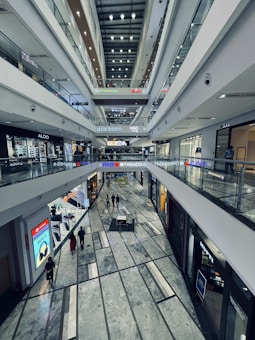 An indoor view of a modern, multi-level shopping mall. The image captures several floors lined with shops and stores, featuring brands like ALDO and M&S. Bright lighting illuminates the corridors, and glass railings separate the different levels. Shoppers walk along the spacious central aisle, and advertisements are visible on digital screens.