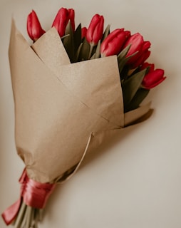 A bouquet of vibrant red tulips wrapped in brown paper with a red ribbon tied around the stems. The flowers are displayed against a neutral background.