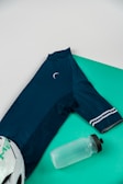 A navy blue cycling jersey with a crescent logo at the neckline, placed on a turquoise background alongside a transparent water bottle with a black lid and a white cycling helmet partially visible.