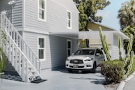 A modern, light gray two-story house with white trim features an outdoor staircase leading to the upper level. A white car is parked under a flat-roofed carport attached to the house. The setting includes several tall green cacti and lush tropical trees and bushes, creating a serene and well-maintained environment.
