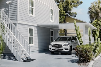 A modern, light gray two-story house with white trim features an outdoor staircase leading to the upper level. A white car is parked under a flat-roofed carport attached to the house. The setting includes several tall green cacti and lush tropical trees and bushes, creating a serene and well-maintained environment.