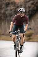 A cyclist wearing a helmet, sunglasses, and a tight-fitting cycling outfit rides a road bike on a paved path. The background is blurred, with a hint of trees and a fence, suggesting motion and speed. The cyclist appears focused and determined.