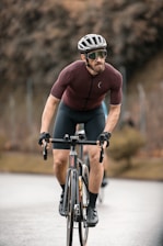 A cyclist wearing a helmet, sunglasses, and a tight-fitting cycling outfit rides a road bike on a paved path. The background is blurred, with a hint of trees and a fence, suggesting motion and speed. The cyclist appears focused and determined.