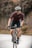 A cyclist wearing a helmet, sunglasses, and a tight-fitting cycling outfit rides a road bike on a paved path. The background is blurred, with a hint of trees and a fence, suggesting motion and speed. The cyclist appears focused and determined.