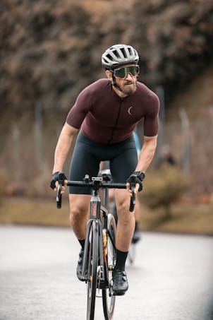 A cyclist wearing a helmet, sunglasses, and a tight-fitting cycling outfit rides a road bike on a paved path. The background is blurred, with a hint of trees and a fence, suggesting motion and speed. The cyclist appears focused and determined.