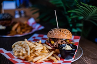 A burger topped with pulled meat, melted cheese, and vegetables sits on a sesame seed bun. Accompanying it is a portion of golden French fries, and a small black cup of mayonnaise is placed near the fries. The food is arranged on a plate lined with a red and white checkered paper, set on a wooden table. In the background, there are blurred images of more food items and green foliage, contributing to a casual dining ambiance.
