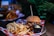 A burger topped with pulled meat, melted cheese, and vegetables sits on a sesame seed bun. Accompanying it is a portion of golden French fries, and a small black cup of mayonnaise is placed near the fries. The food is arranged on a plate lined with a red and white checkered paper, set on a wooden table. In the background, there are blurred images of more food items and green foliage, contributing to a casual dining ambiance.