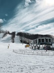 A snowy ski resort scene with people skiing and a ski lift station visible. The landscape is covered in snow, with bare trees lining the slope. A partially cloudy sky and bright sunlight create a serene winter atmosphere.