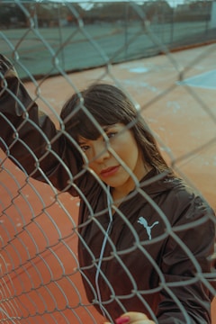 A person wearing a dark sports jacket is positioned behind a chain-link fence on a tennis court. The surface of the court is orange, and the person gazes directly at the camera, resting one arm on the fence. The setting appears to be outdoors with elements of urban decay.