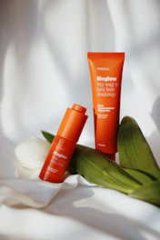 Two orange cosmetic products labeled 'Bioglow' are placed on a white, slightly wrinkled fabric with fresh green leaves and a white tulip resting beside them. The packaging is vibrant and stands out against the soft background.