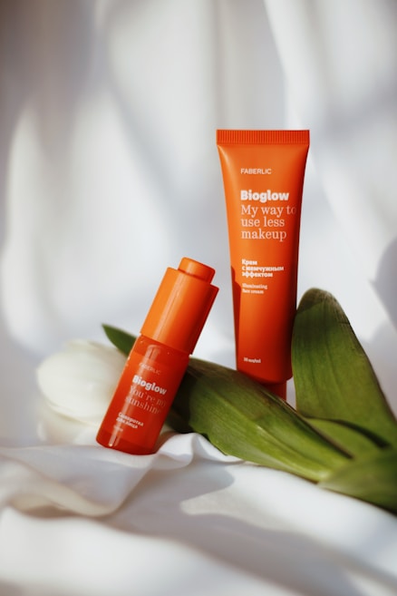 Two orange cosmetic products labeled 'Bioglow' are placed on a white, slightly wrinkled fabric with fresh green leaves and a white tulip resting beside them. The packaging is vibrant and stands out against the soft background.