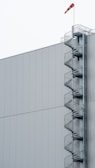 A large, plain industrial building with a spiral staircase attached to the right side. The staircase extends vertically and is made of metal, with protective railings. At the top, a red and white windsock indicates wind direction.