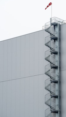 A large, plain industrial building with a spiral staircase attached to the right side. The staircase extends vertically and is made of metal, with protective railings. At the top, a red and white windsock indicates wind direction.