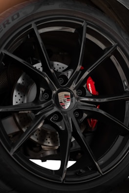 A detailed look at a car wheel featuring a glossy black alloy rim with a branded emblem at the center. The disc brake system is visible behind the rim, including drilled rotors for performance braking. The red brake caliper adds a vibrant accent, contrasting with the black wheel.
