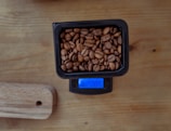 A small rectangular black container filled with coffee beans is placed on a digital scale. The blue screen of the scale is illuminated, showing a measurement. The setup is on a wooden surface, next to a wooden cutting board with a hole in its corner.