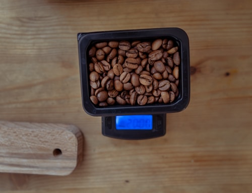 A small rectangular black container filled with coffee beans is placed on a digital scale. The blue screen of the scale is illuminated, showing a measurement. The setup is on a wooden surface, next to a wooden cutting board with a hole in its corner.