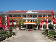 A large building with three floors and a red tile roof, surrounded by red flags on poles lining a walkway. The facade displays vibrant banners with text in a foreign script. The entrance is decorated with potted plants and a small staircase, leading to a path flanked by green shrubs and trees.