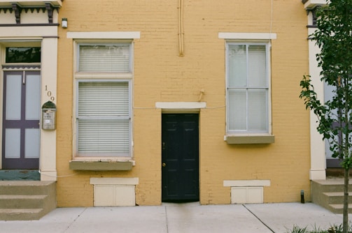 A brick building with a mustard-yellow facade features a small black door in the center, flanked by two large windows with white frames and blinds. The structure has steps leading up to a purple and gray door on the left with the number '109' displayed. A small mailbox is mounted near the door, and a tree with green leaves is partially visible on the right side.