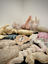 A collection of large, soft sculptures resembling oversized plush body parts, such as hands and arms, are arranged on the floor and against white walls. These sculptures are made from various fabrics in neutral and pastel shades. The artwork creates a surreal and abstract setting.